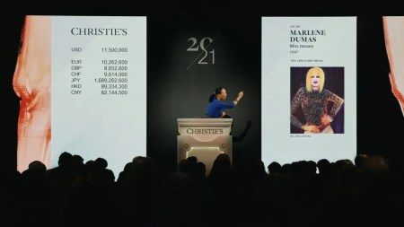 An auctioneer at a podium beside an image of a painting showing a woman with one hand bent at her waist.