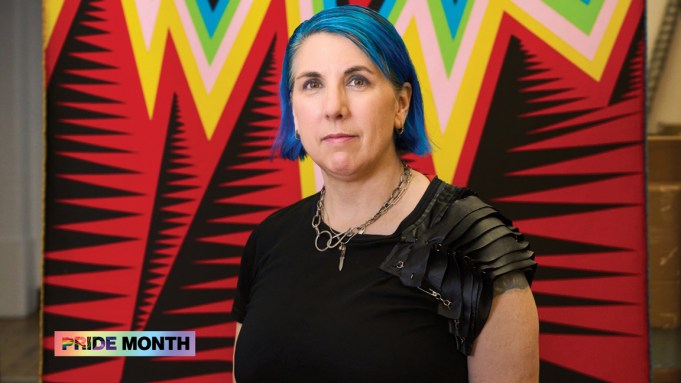 A woman with dyed-blue hair standing in front of a weaving featuring zigzagging lines in various colors.