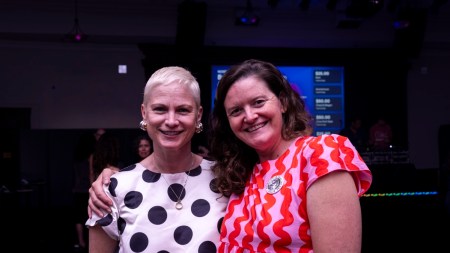 A woman with a blonde pixie cut in a black and white polka dot dress, stands next to a woman with long curly brown hair, and a pink dress with red squiggles.