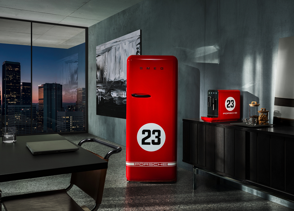 A modern, stylish interior with a large window revealing a cityscape at dusk. The room features sleek dark furniture and minimalist decor. Prominently displayed are a red SMEG refrigerator and a red coffee machine, each marked with a white circle containing the number "23" and "PORSCHE" logo. The refrigerator has the brand name "SMEG" in bold letters at the top. Behind it, a black and white abstract painting adds contrast to the scene. The design emphasizes bold colors, clean lines, and strong visual contrast, ensuring legibility and accessibility for users with visual impairments.